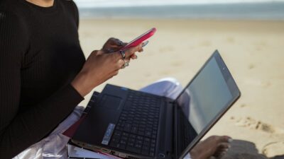 Person multitasking with a laptop and smartphone by the beach, embracing remote work lifestyle.